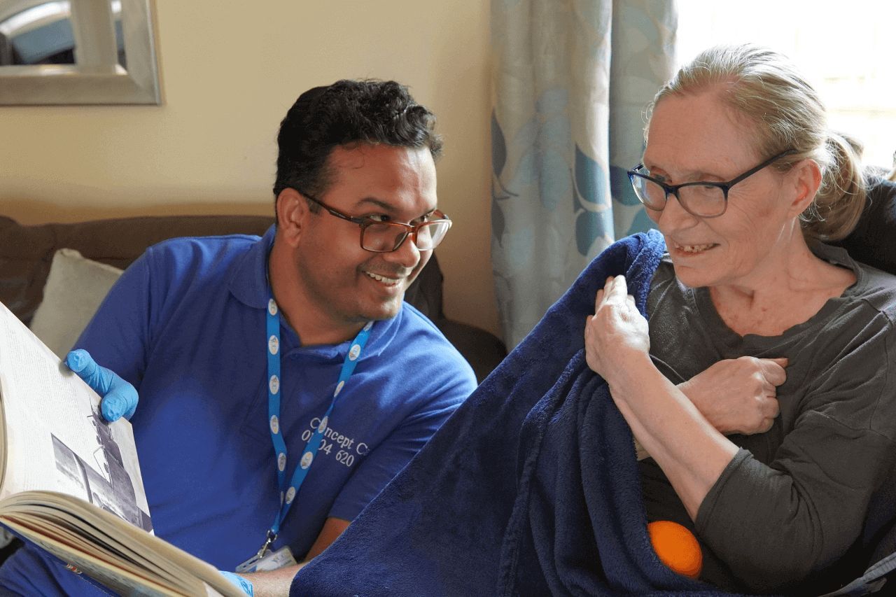 A caregiver from Concept Care Community Services sitting with an elderly woman, both smiling warmly while reading a book together.