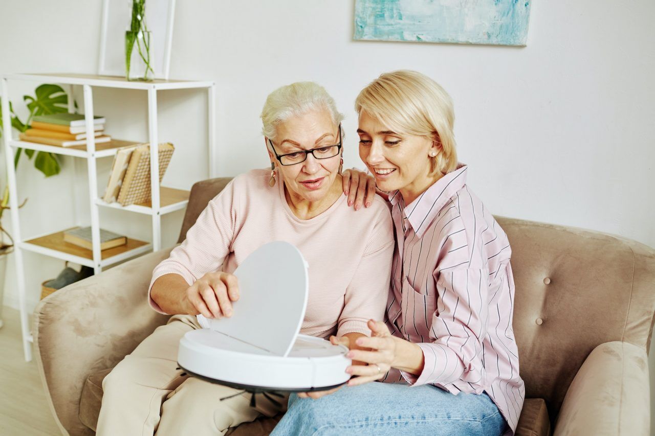 A senior woman sitting on a couch holding a smart vacuum cleaner, with a younger caregiver by her side, both smiling as they look at the device.