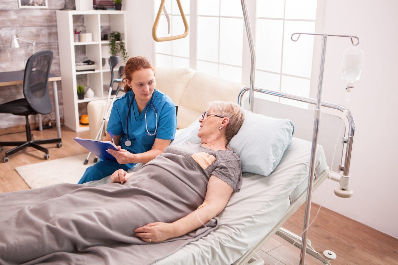 A senior woman lying in a nursing home bed, being comforted by a caregiver who is sitting beside her and holding her hand.