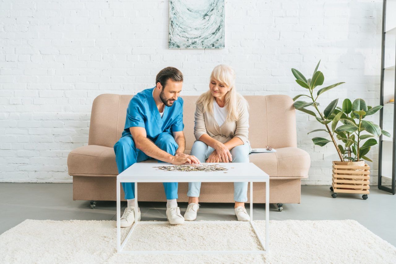 A senior woman and a young caregiver sitting on a couch, smiling and working together on a jigsaw puzzle.